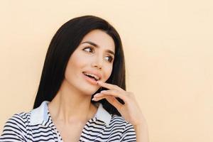 complacida y encantadora mujer joven con cabello oscuro, se ve feliz y alegremente a un lado, posa contra un fondo claro, vestida informalmente, tiene la piel limpia, manicura, mantiene la mano cerca de la boca, modelos en el estudio foto