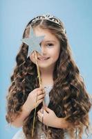 Isolated image of satisfied beautiful curly woman with blue eyes, covers face with magic wand, has long curly hair, poses over blue background, gentle smile on face, makes wish, dreams about something photo