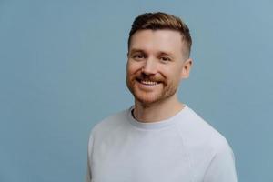 Handsome cheerful bearded man smiles gladfully dressed in casual white t shirt being in good mood poses against blue background with copy space. Glad European guy with stubble stands indoor. photo