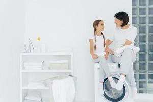 Caring mother talks with daughter, pose on washing machine, surrounded with white linen, do washing together, being in laundry room at home. Happy adult housewife thanks adorable girl for help photo