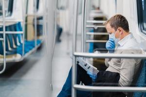 Prohibition of free movement. Man in formal clothes, coughs and has symptoms of coronavirus, wears protective medical mask and gloves, reads newspaper, poses in subway or metro, commutes to work photo