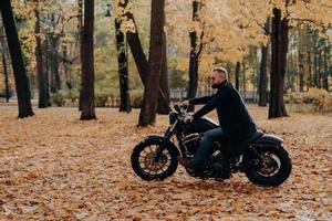 Outdoor image of male motorcyclist poses on fast motorbike, wears shades, black coat, enjoys ride in autumn park, breathes fresh air, admires nature during sunny day. Active bearded man outside photo