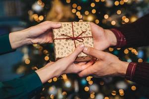 Wrapped box in womans and mans hands. Affectionate husband congratulates his wife with New Year, presents her holiday gift box, stand against decorated Christmas tree. Holiday concept photo
