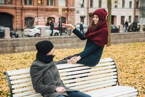 Good looking female wers knitted hat and scarf, throws yellow leaves in air, holds boyfriends hand, have fun together as sit on bench together. People, relationshp concept photo