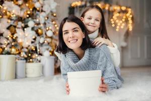 toma interior de madre e hija que se divierten juntas, comparten regalos, están en una habitación decorada con guirnaldas y árboles de navidad, tienen expresiones alegres, disfrutan los fines de semana y las vacaciones de invierno foto