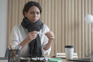 Unhealthy young hispanic business woman wearing scarf around neck holding thermometer photo