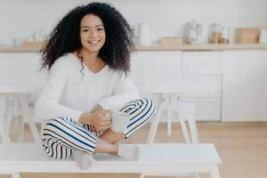 Smiling relaxed African American female sits crossed legs on bench against kitchen interior, wears white sweater and striped pants, drinks hot beverage, enjoys domestic atmosphere. Coffee time photo