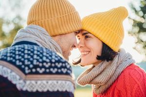 Adorable brunette female and her boyfriend stand closely to each other, look at eyes, smile happily as feel love and support. Good looking woman with scarf and hat has date with young attractive man photo