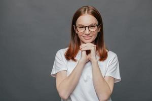 una dama alegre y de aspecto agradable mantiene ambas manos debajo de la barbilla, sonríe positivamente a la cámara, usa anteojos ópticos, camiseta informal, disfruta del tiempo libre en buena compañía, aislada en un fondo gris foto