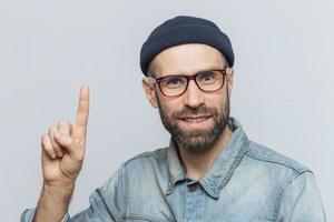 People, emotions and reaction concept. Handsome bearded guy in trendy eyewear, has pleased expression, raises fore finger as remembers something positive, isolated over grey studio background photo