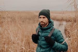 People and tourism concept. Pensive unshaven handsome young man with rucksack, dressed in stylish clothes, focused into distance, poses against outdoor field background. Lifestyle. Adventure photo