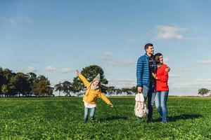 Playful little child runs on green meadow, plays near parents, who embrace each other, look into distance with thoughtful expressions. Family has walk or stroll in countryside, rest after noise city photo