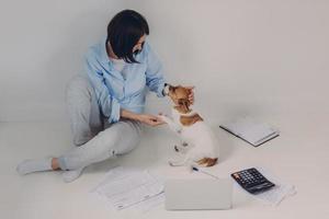 Brunette woman dressed casually, studies income report, sits on floor with laptop computer, paper documents, calculator, notepad, has fun with her favourite pet, isolated over white background photo