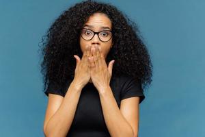 Photo of stupefied young female covers mouth, has bated breath, bugged eyes, Afro hair, cannot believe in shocking news, wears casual black t shirt, stands over blue background. Omg concept.