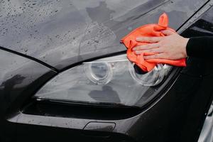 Mans hand with rag wiping cars headlamp. Wet black automobile cleanes at car wash. Auto cleaning and service concept photo