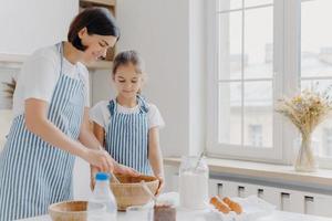 Photo of brunette woman in striped apron, mixes ingredients with beater, shows small daughter how to cook, stand at kitchen near table with fresh products. Mother and child busy preparing meal