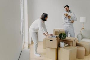 Busy woman tries to find information in laptop computer, buys furniture online, man stands with dog on hands, smiles and talks to wife, surrounded with pile of cardboard boxes have to unpack property photo