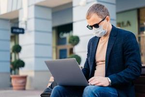 Concentrated businessman poses outdoor near bus station, uses laptop computer for distance work, wears protective mask to avoid coronavirus. People, technology, travel and virus outbreak concept