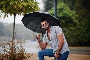 Fashionable tall arab beard man wear on shirt, jeans and sunglasses with umbrella posed at rain on park square. photo