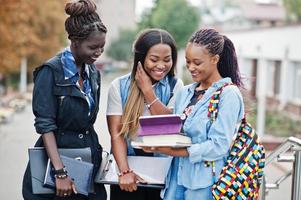 Three african students female posed with backpacks and school items on yard of university and look at tablet. photo