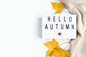 Hola otoño. firmar con la inscripción, suéter de punto y hojas caídas sobre un fondo blanco. comienzo de la temporada de otoño. endecha plana foto