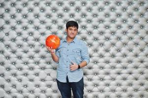 Stylish asian man in jeans shirt standing with bowling ball at hand against silver wall background. photo