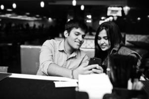 Stylish asian couple wear on jeans sitting on chair at bar in club and look on phone. photo