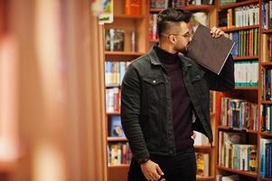 Tall smart arab student man, wear on black jeans jacket and eyeglasses, at library with book at hands. photo