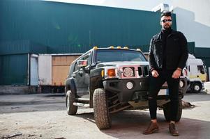 El hombre árabe de barba rica en moda usa una chaqueta negra de jeans y gafas de sol posadas contra un gran auto todoterreno negro. chico modelo árabe elegante, exitoso y de moda. foto