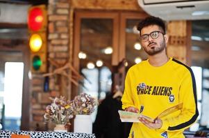 hombre asiático con anteojos y sudadera amarilla posando en un café interior con semáforo. foto