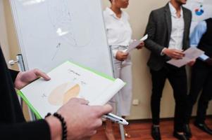 Mixed race business coach presenting report standing near whiteboard pointing on sales statistic shown on diagram and chart teach diverse company members gathered together in conference room. photo