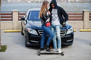 Cool pareja multirracial se abraza junto con longboard contra elegante coche deportivo negro. foto