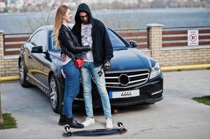 Cool pareja multirracial se abraza junto con longboard contra elegante coche deportivo negro. foto