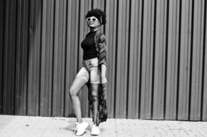 African woman with afro hair, in jeans shorts and white sunglasses posed against green steel wall. photo