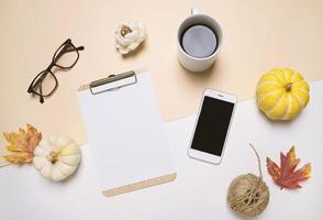 Creative flat lay of workspace desk in autumn style with blank clipboard, smartphone, eyeglasses, pumpkins, coffee and autumn leaves with copy space background, minimal style photo
