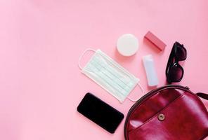 Flat lay of red leather woman bag open out with cosmetics, accessories, smartphone and mask with alcohol gel sanitizer on pink background, copy space and prevent of Covid-19 and Coronavirus concept photo