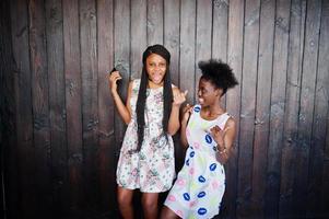 Two black african girlfriends at summer dresses posed against dark wooden background and having fun. photo