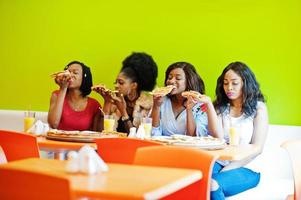 Four young african girls in bright colored restaurant eating pizza slices in hands. photo