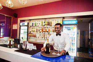 African american bartender at bar holding champagne with glasses on tray. photo