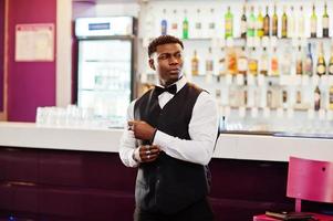 joven apuesto hombre africano vestido con camisa blanca, chaleco negro y corbata de moño posado contra el mostrador del bar en el club nocturno. foto