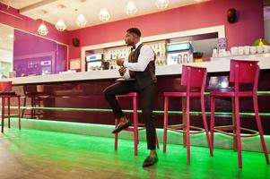 Young handsome african man wearing white shirt, black vest and bow tie with wallet purse posed against bar counter at night club. photo