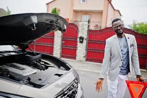 Stylish and rich african american man stand in front of a broken suv car needs assistance looking under opened hood and hold e,ergency triangle. photo