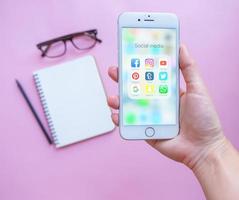 Hand holding Apple iPhone 6s with group of Popular Social networks icons showing on  screen with blank notebook and eyeglasses on pink background, Social media are most popular tool for communication. photo
