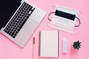 Top view flat lay of workspace desk with laptop, blank notebook, medical masks and alcohol gel on bright pink background photo