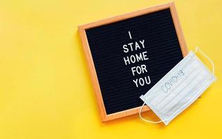 Flat lay of notice board with message I stay home for you and medical mask written covid-19 on yellow background with copy space