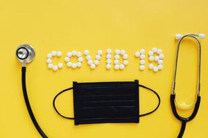 Flat lay of medicine pills shape as word covid 19 with black mask and stethoscope on yellow background, health care concept and prevent the spread of pandemic Covid-19 and Coronavirus photo