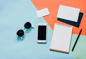 Creative flat lay style workspace desk with smartphone, blank envelope, sunglasses and blank notebook on modern colorful background photo