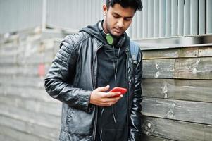 hombre asiático elegante e informal con chaqueta de cuero negro, auriculares con teléfono móvil rojo en las manos posado en la calle. foto