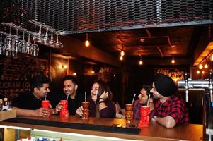 Group of indian friends having fun and rest at night club, drinking cocktails near bar counter. photo