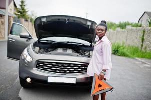 Young sad african woman stand with emergency triangle against car with popped hood. Transportation, vehicles problems and breakdowns concept. photo